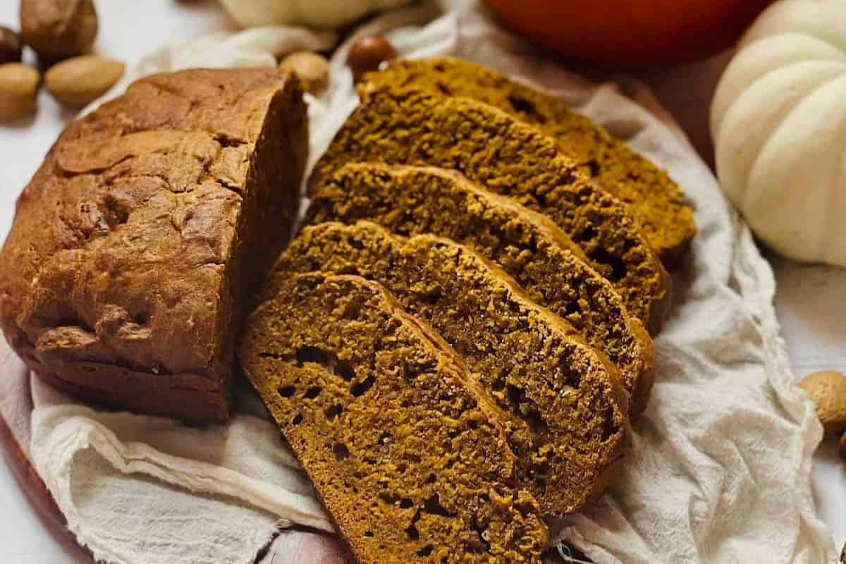 A loaf of pumpkin bread, a must-try among quick breads, sits partially sliced with three slices fanned out on a cloth, surrounded by whole nuts and decorative white pumpkins.