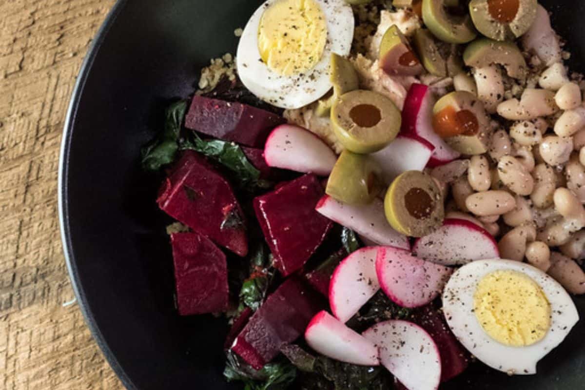A close-up of a quinoa bowl filled with various ingredients including diced cooked beets and their greens, sliced radishes, hard-boiled egg halves, green olives, and white beans, all in a black bowl on a wooden surface.