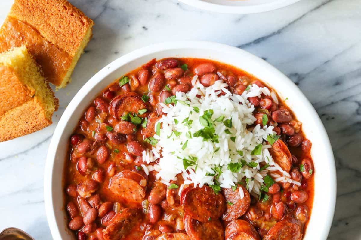 A bowl of red beans and sausage made with canned beans, topped with white rice and herbs, served next to pieces of cornbread on a marble surface—one of those delicious dishes perfect for easy recipes at home.