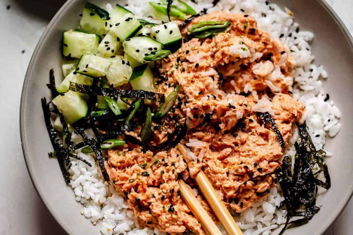 A bowl of white rice topped with spicy salmon, sliced cucumbers, seaweed strips, and black sesame seeds—an easy dinner idea that can also be made with canned tuna for a twist on classic tuna recipes. Chopsticks rest on the side.