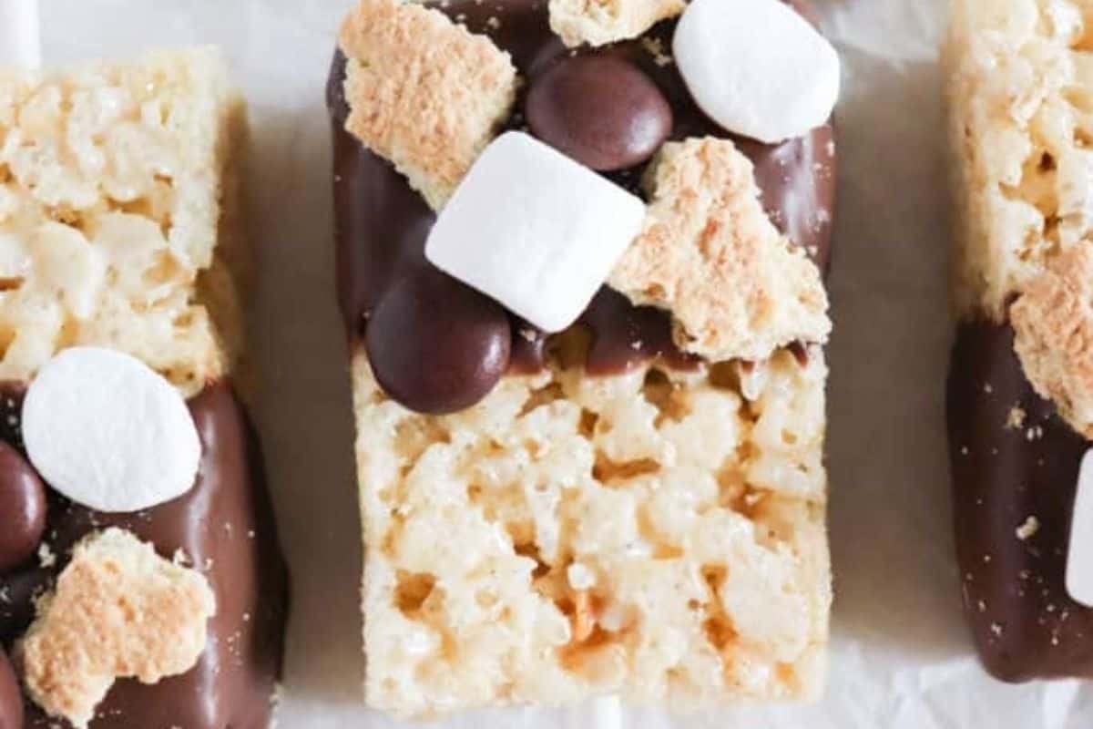 A top-down close-up view of two s'mores Rice Krispie treats, likely on sticks, partially dipped in chocolate and topped with mini marshmallows, chocolate chips, and crumbled graham crackers. One treat is in focus in the center, showcasing its crispy texture.