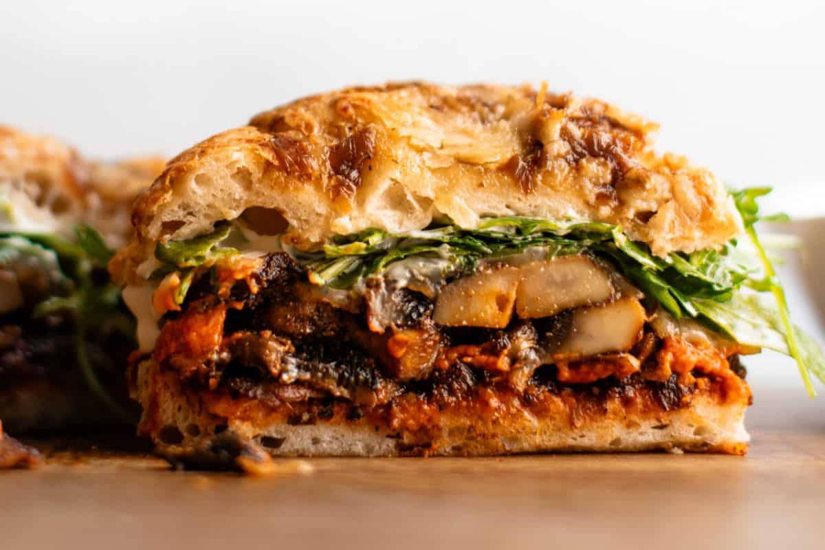 A close-up of a gourmet sandwich with grilled mushrooms, leafy greens, red sauce, and a crusty bread bun on a wooden surface—perfect for lovers of tasty sandwiches.