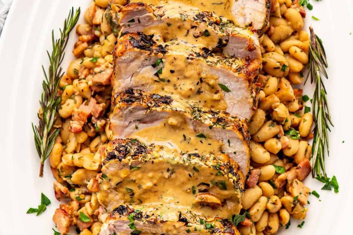 Sliced delicious pork roast topped with sauce, served on a bed of white beans and garnished with rosemary sprigs—a standout among pork roast recipes.