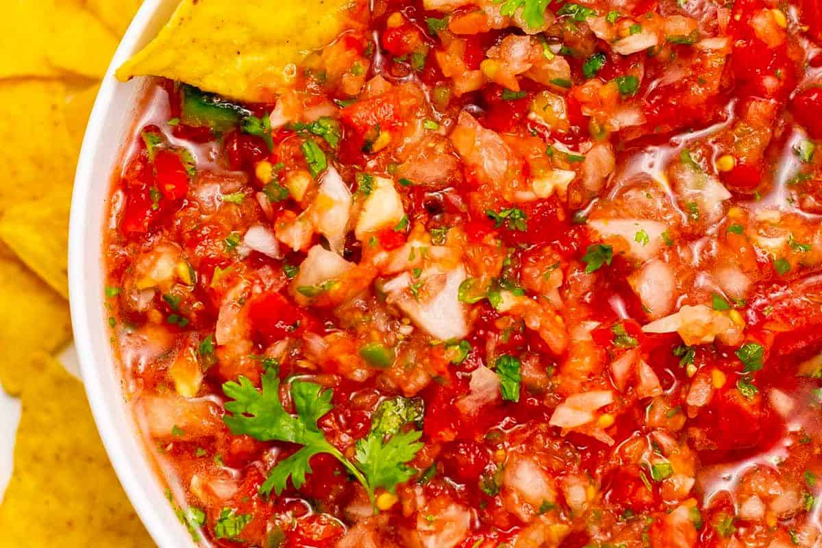 A close-up of fresh salsa in a white bowl, showing diced tomatoes, onions, cilantro, green peppers, and a hint of fresh lime—perfect for those who love vibrant recipes and inspired cooking. Tortilla chips sit ready on the side.
