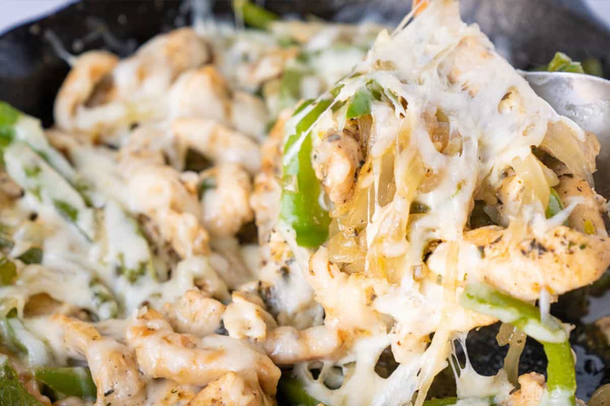 A close-up of a skillet filled with cooked chicken pieces, green bell peppers, and onions, all covered in melted, stringy white cheese. A spoonful is lifting some of the mixture.