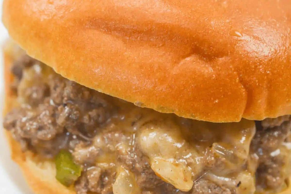 A close-up of a sloppy joe sandwich on a golden-brown bun, showing the rich, meaty, and cheesy filling with bits of onion and green pepper.