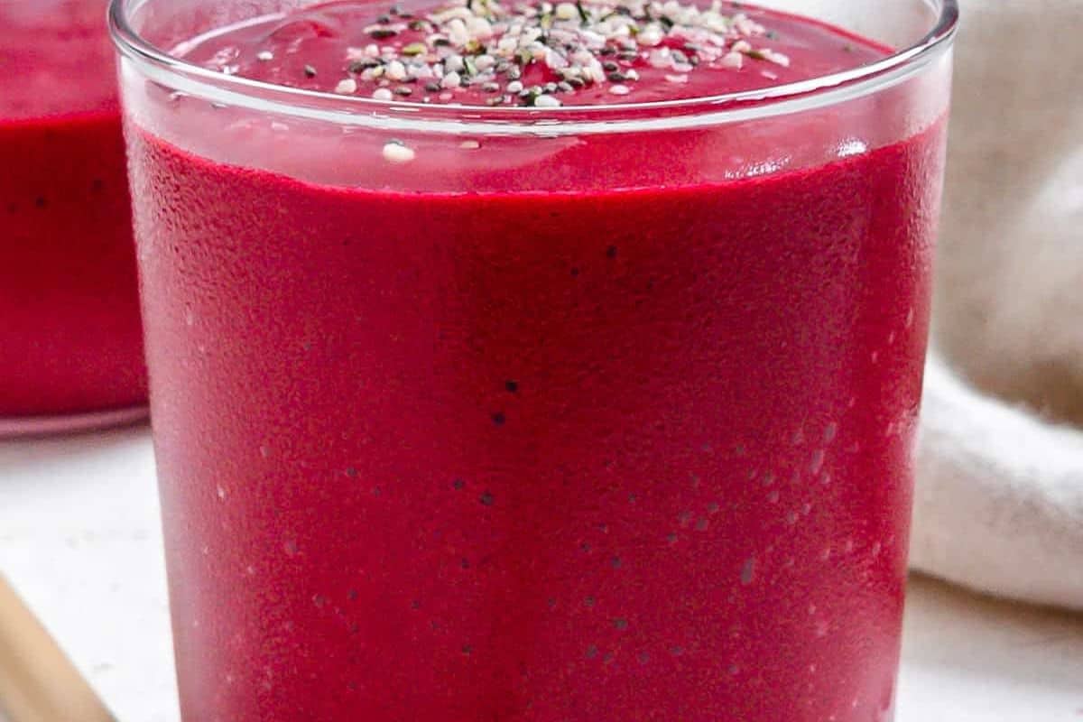 A vibrant red smoothie in a clear glass, topped with a sprinkle of multi-colored seeds (possibly hemp seeds, chia seeds, and poppy seeds).