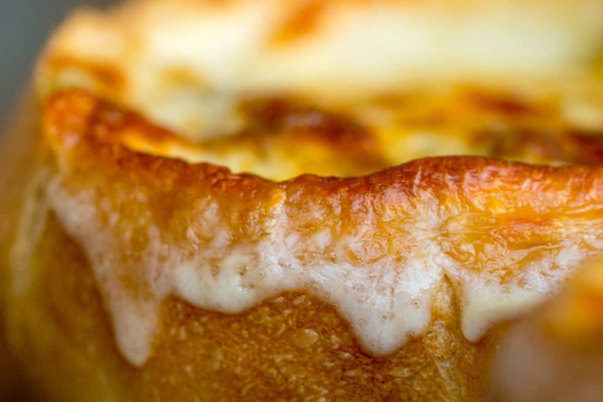 A very close-up, shallow depth of field shot of the rim of a bowl of French onion soup, showing melted, bubbling golden-brown cheese overflowing and dripping down the side of the bread or crouton.