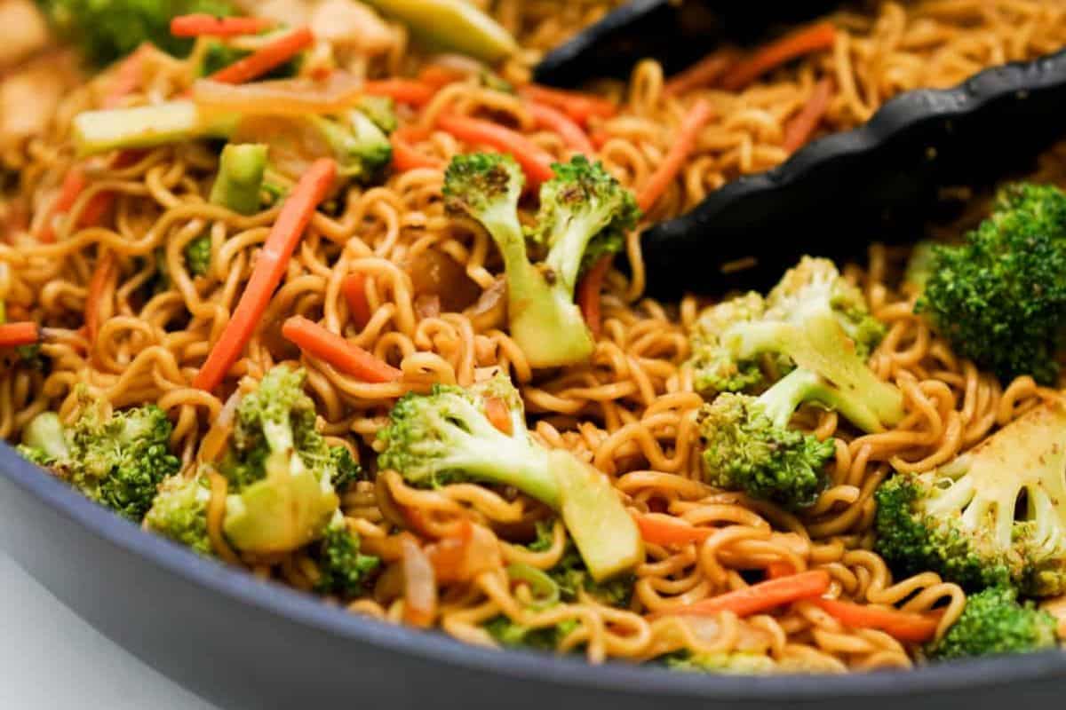 Close-up of a pan filled with flavor packed ramen, broccoli florets, and julienned carrots, with a pair of black tongs resting on top.