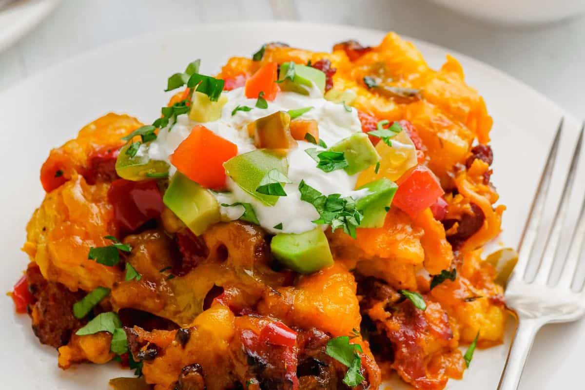 A serving of cheesy casserole—perfect for busy mornings—topped with sour cream, diced avocado, tomatoes, and chopped herbs on a white plate with a fork. This must try dish brings comfort to slow cooker breakfasts.