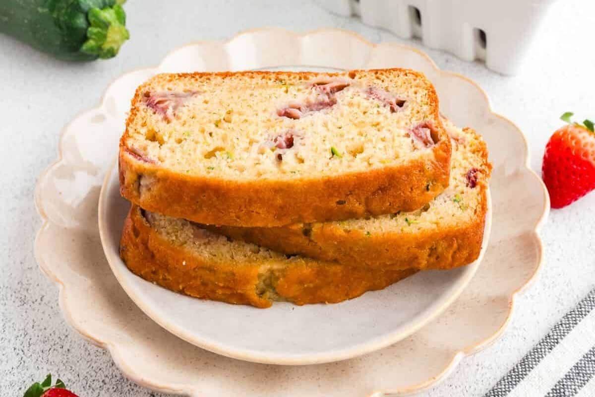 Two slices of strawberry zucchini bread are stacked on a white plate, with a fresh zucchini and strawberries nearby—a must-try for fans of quick breads you can bake anytime.