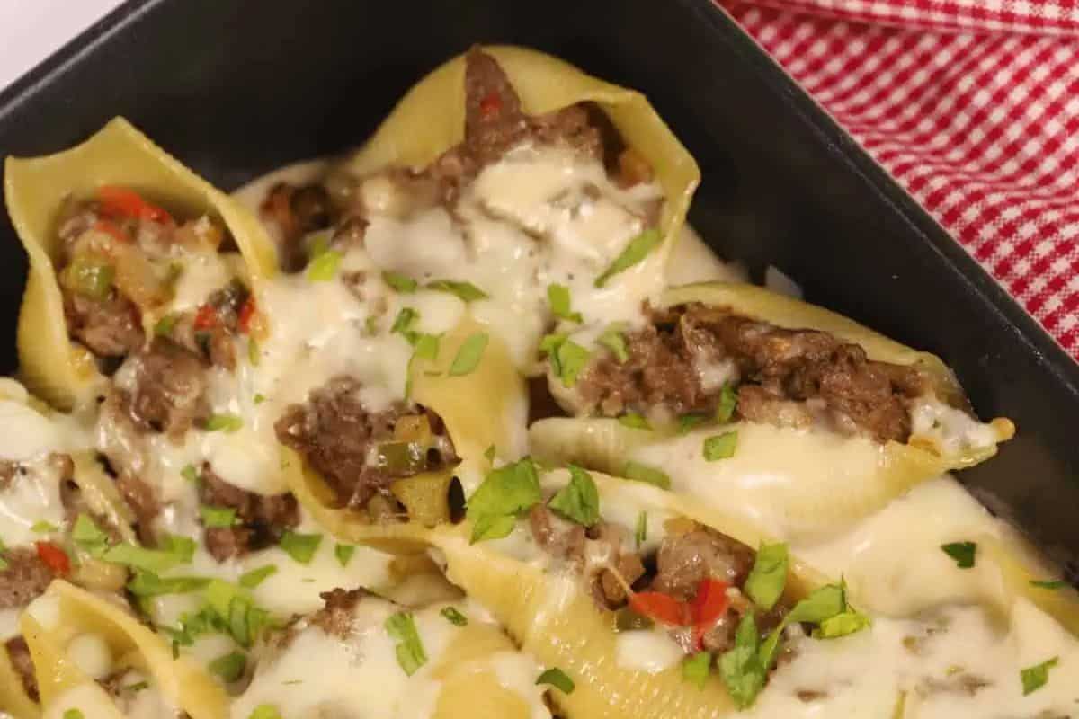 Several large pasta shells generously stuffed with a meat and vegetable mixture, covered in a creamy white cheese sauce, and sprinkled with fresh green herbs, resting in a black baking dish with a red and white checkered cloth in the background.