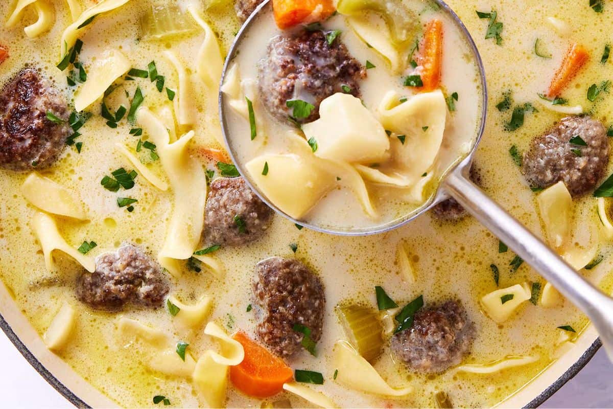 A ladle serving creamy, comforting beef soup with meatballs, egg noodles, carrots, celery, and parsley from a pot—a delicious addition to your favorite dinner recipes.