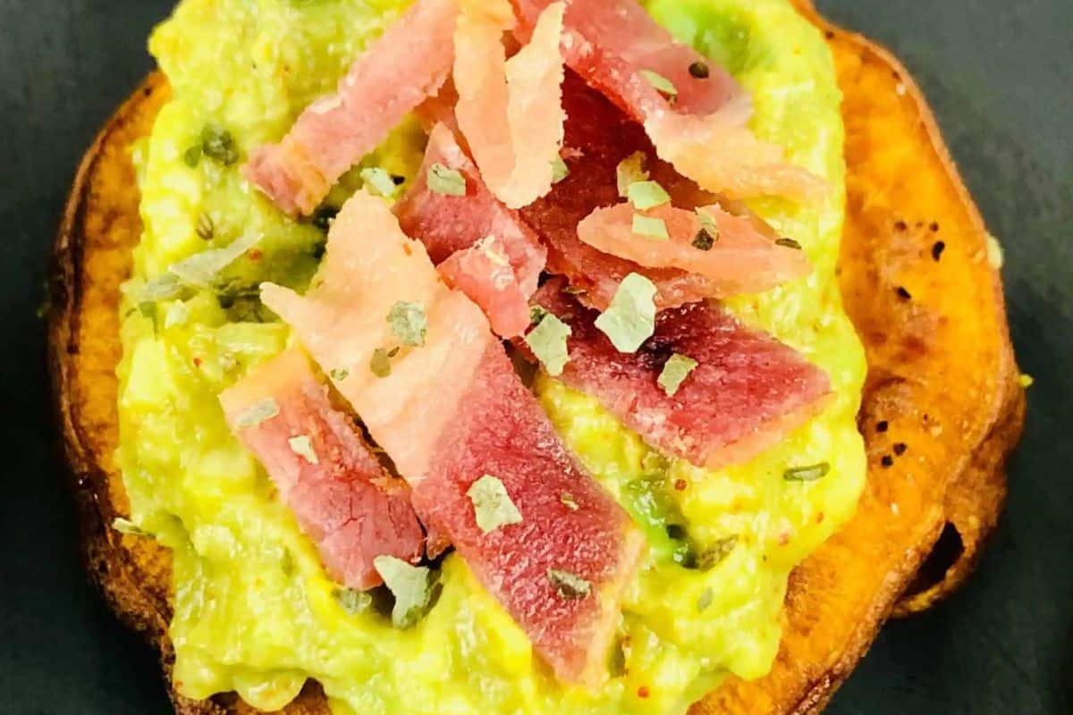 A slice of roasted sweet potato topped with mashed avocado and strips of cured meat, garnished with herbs—one of the tastiest cold appetizer ideas for your next gathering.
