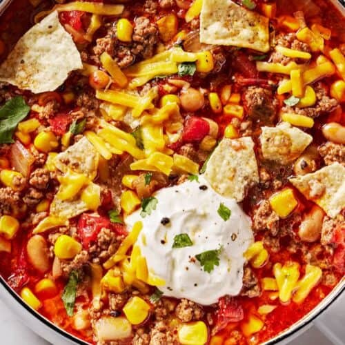A comforting bowl of taco soup, perfect for dinner, topped with shredded cheese, tortilla chips, corn, ground beef, beans, tomatoes, and a dollop of sour cream with herbs—one of the coziest beef soup recipes to enjoy.