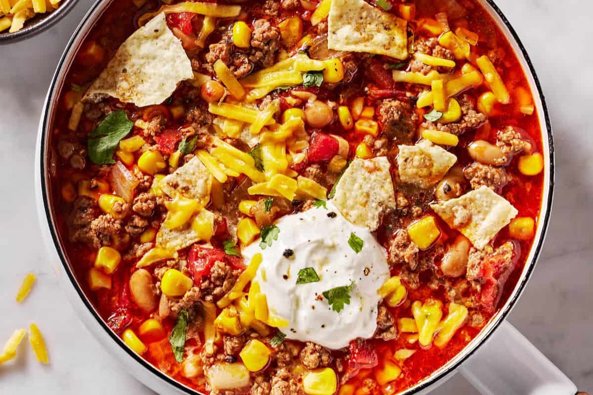 A comforting bowl of taco soup, perfect for dinner, topped with shredded cheese, tortilla chips, corn, ground beef, beans, tomatoes, and a dollop of sour cream with herbs—one of the coziest beef soup recipes to enjoy.