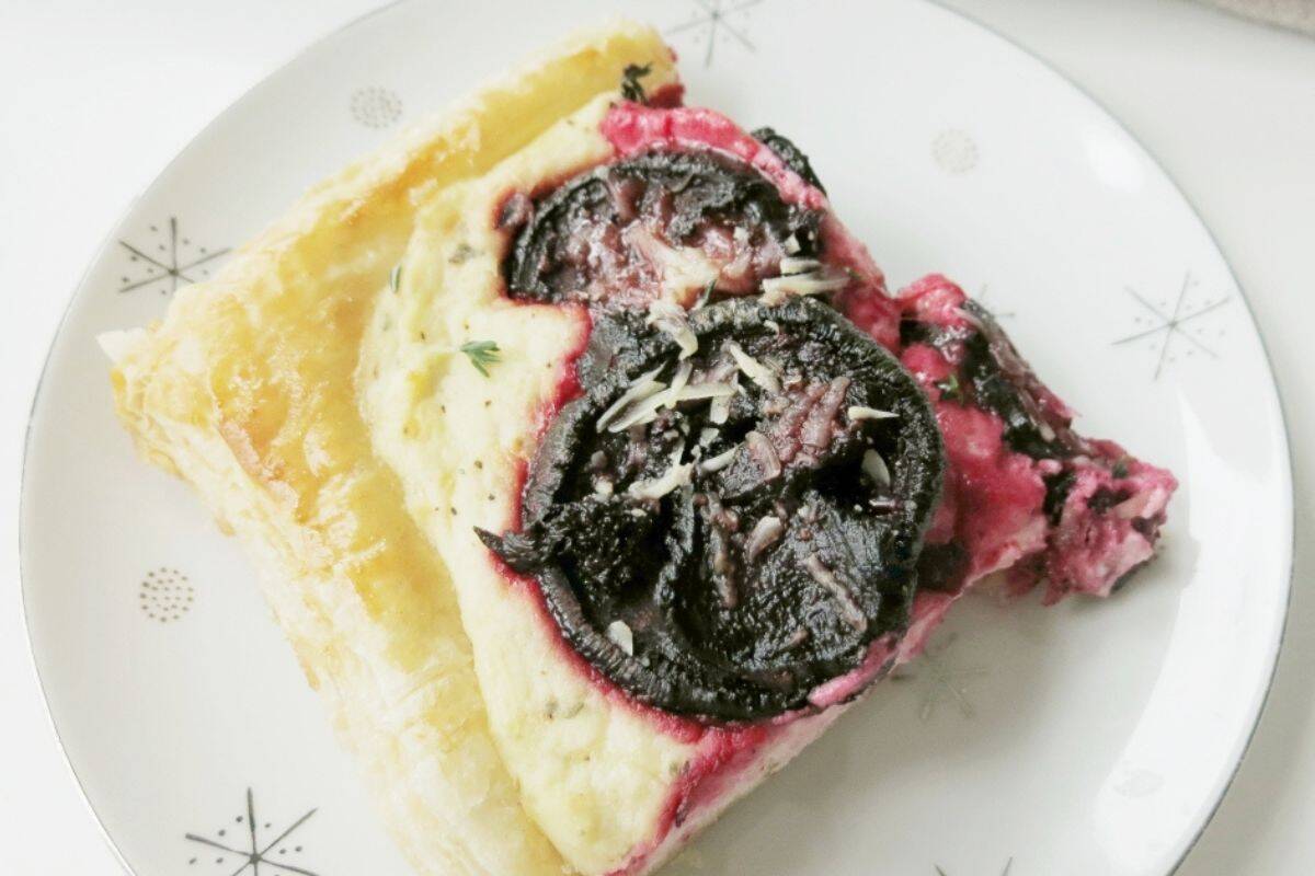  A slice of a savory tart on a white plate with a subtle starburst pattern. The tart has a flaky golden crust, a creamy white filling, and is topped with dark, sliced roasted beets and a sprinkle of grated cheese or herbs.