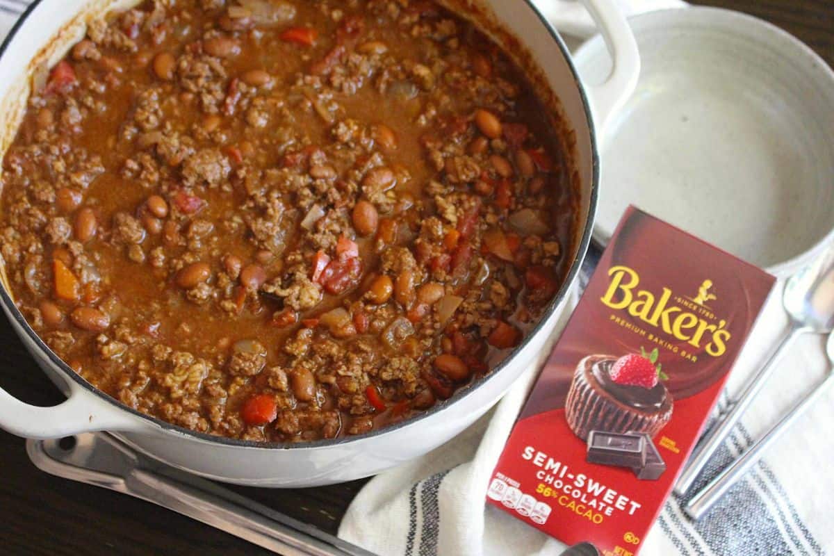 A pot of chili with ground beef and canned beans sits next to a box of Baker’s semi-sweet chocolate and two empty bowls with spoons, ready for serving delicious dishes inspired by classic recipes.