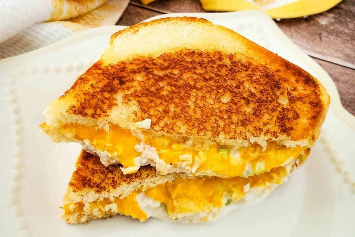 A grilled cheese sandwich cut in half, stacked on a white plate, reveals melted cheese and a savory canned tuna dinner filling inside golden toasted bread—perfect for easy tuna meals.