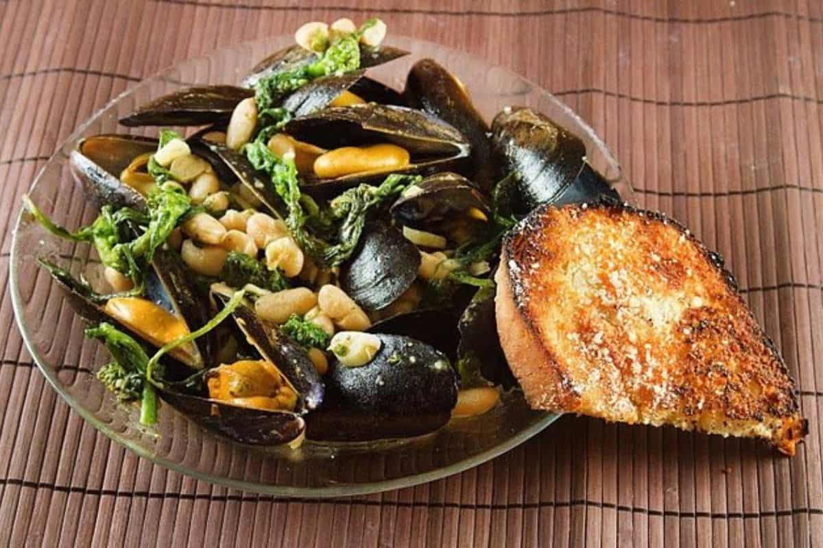 A glass plate with cooked mussels, canned beans, greens, and a slice of toasted bread, placed on a brown bamboo mat.