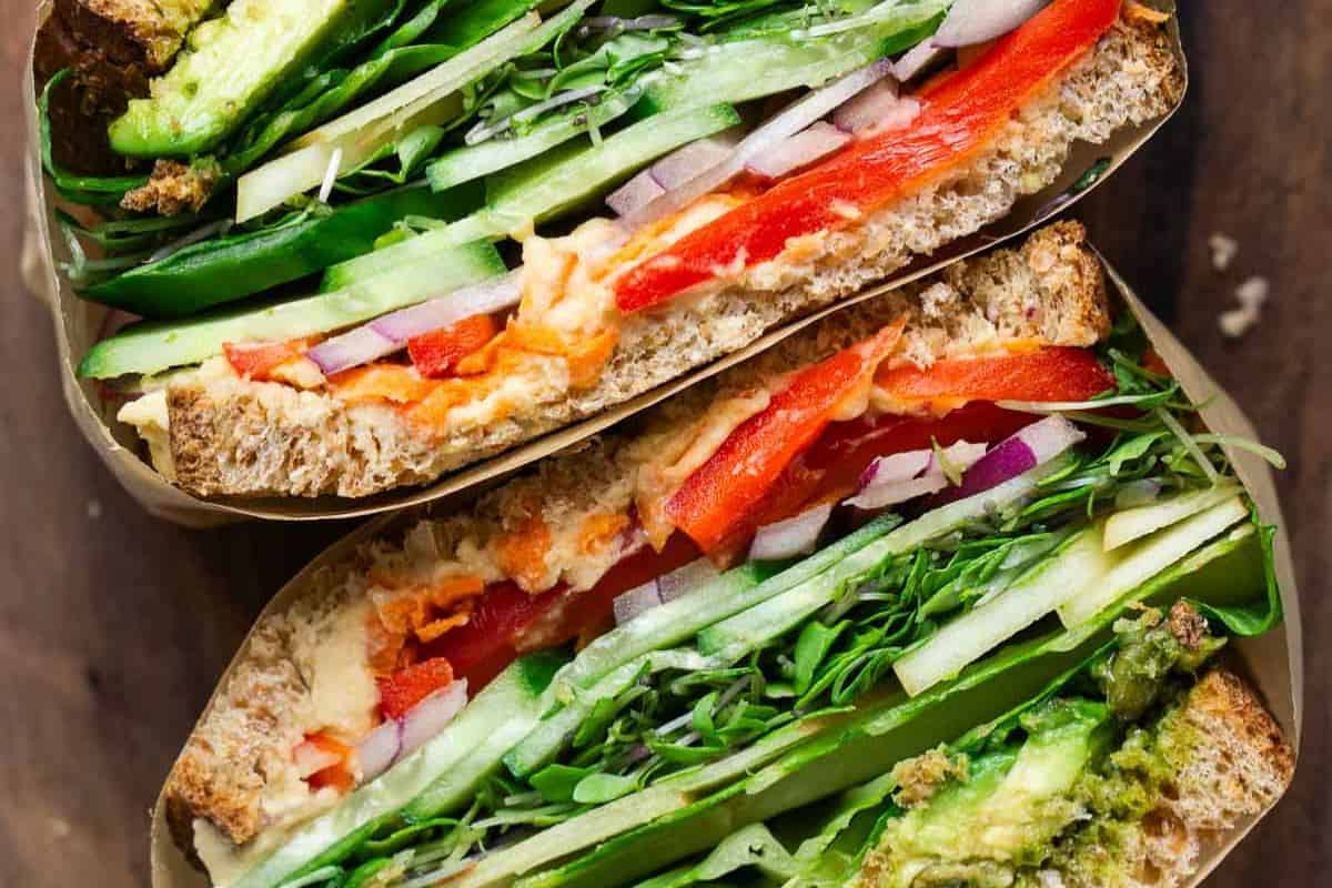 A close-up of two stacked gourmet sandwiches on whole grain bread, filled with lettuce, cucumber, red bell pepper, onion, sprouts, and spreads—these visually appealing sandwiches are bursting with fresh flavors.