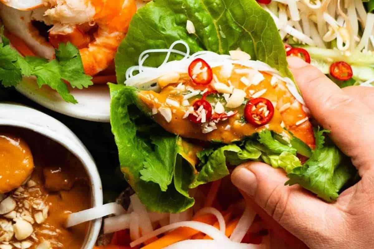 A hand holding a vibrant Vietnamese lettuce wrap filled with shrimp, rice noodles, fresh herbs, and a generous dollop of peanut sauce, with more ingredients in the background.