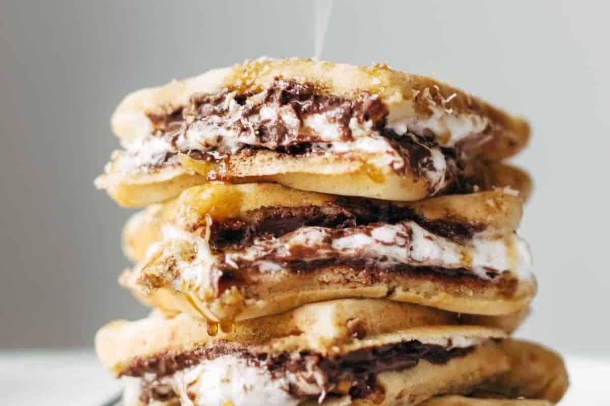  A tall stack of three s'mores waffle sandwiches, seen from a slightly low angle. Each sandwich features golden-brown waffles filled with melted chocolate and gooey white marshmallows. A drizzle of syrup or honey is falling onto the top waffle and dripping down the sides.