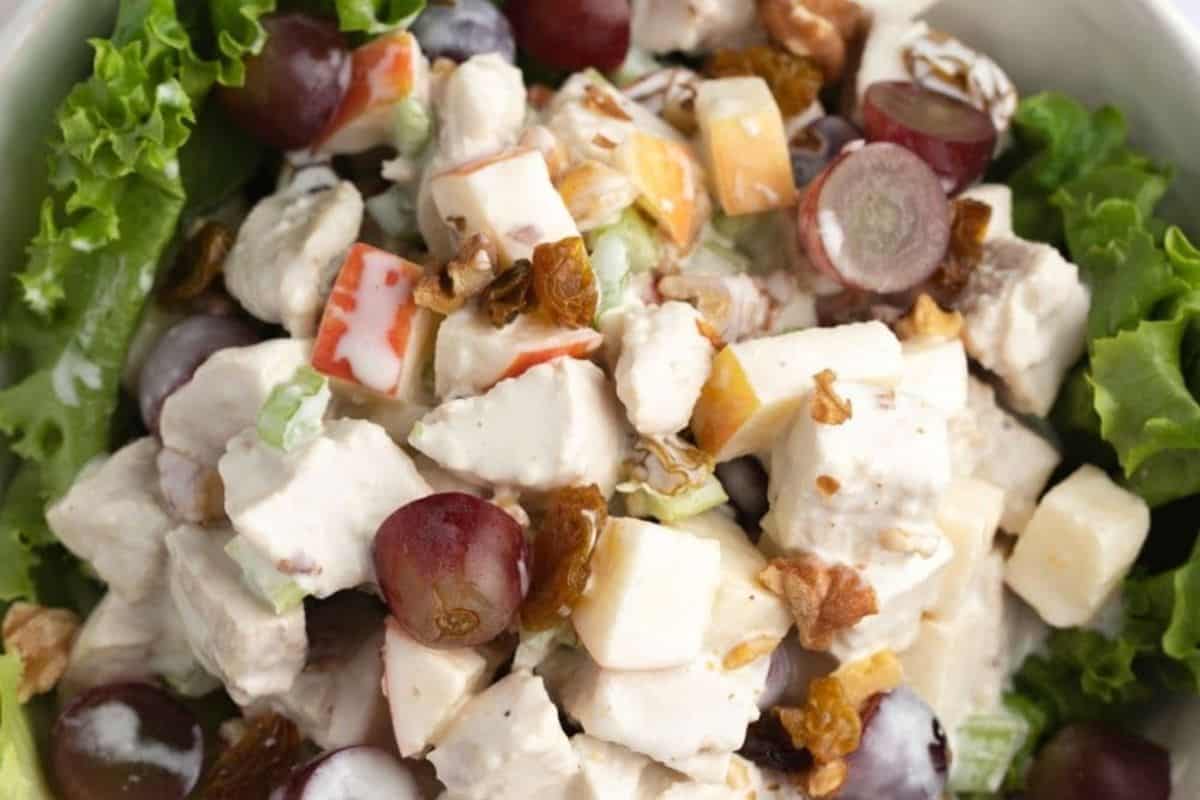 A close-up of a Waldorf salad with chunks of chicken, red grapes, celery, walnuts, and golden raisins, all mixed in a creamy dressing and served on a bed of green lettuce.