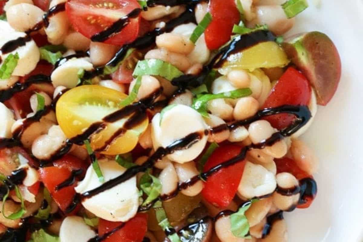  A close-up of a white bean caprese salad with white beans, cherry tomatoes, fresh mozzarella balls, and basil, drizzled with a dark balsamic glaze.