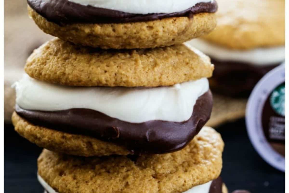 A stack of three s'mores whoopie pies on a dark surface, with a blurred background showing burlap and a coffee pod. Each whoopie pie consists of two soft, light brown cookies with a thick filling of dark chocolate and white marshmallow cream in between.