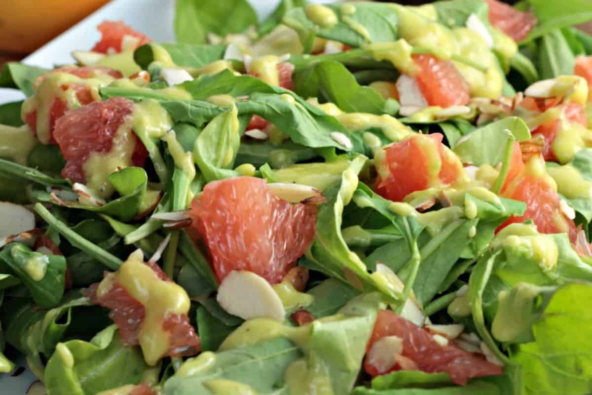 A close-up of a fresh salad with arugula, juicy grapefruit segments, sliced almonds, and a creamy yellow dressing—perfect for fans of refreshing grapefruit recipes.