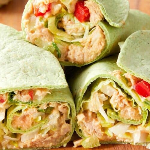 Three spinach wraps filled with tuna salad, lettuce, diced tomatoes, and onions are stacked on a wooden surface—a delicious example of easy tuna meals.