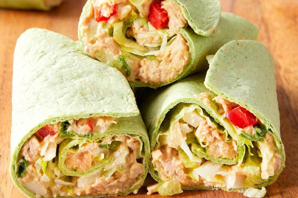 Three spinach wraps filled with tuna salad, lettuce, diced tomatoes, and onions are stacked on a wooden surface—a delicious example of easy tuna meals.