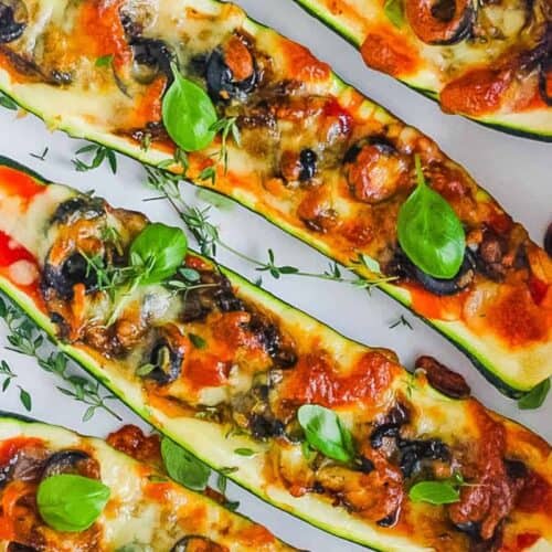 Four baked zucchini halves filled with melted cheese, tomato sauce, olives, and herbs, garnished with fresh basil and thyme—an inviting dish perfect for party food recipes, beautifully arranged in a white baking dish.