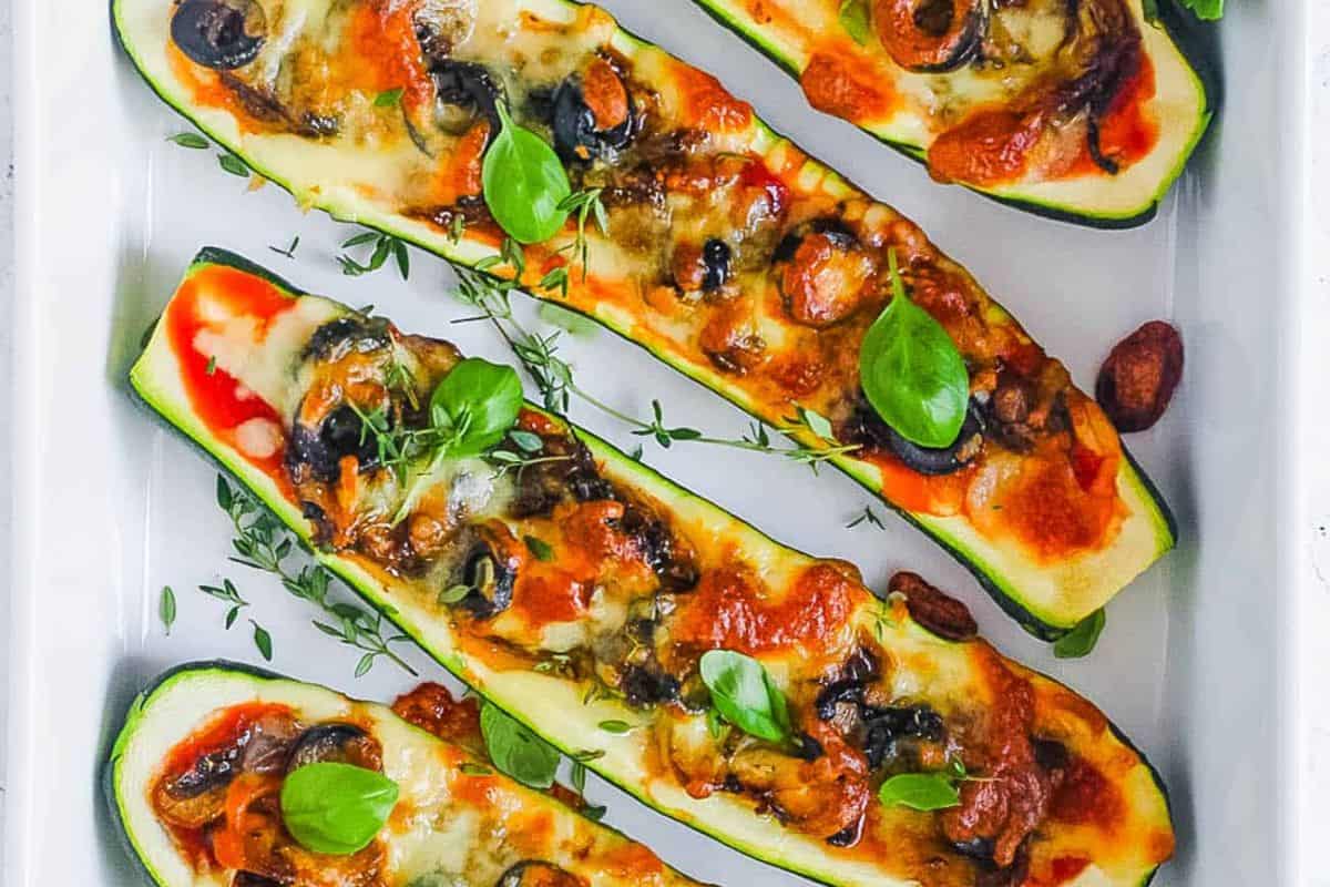 Four baked zucchini halves filled with melted cheese, tomato sauce, olives, and herbs, garnished with fresh basil and thyme—an inviting dish perfect for party food recipes, beautifully arranged in a white baking dish.