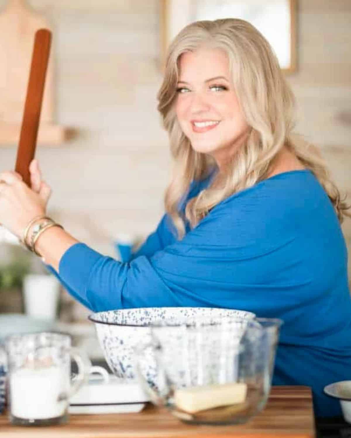 A woman in a blue top smiles while holding a rolling pin in a kitchen filled with mixing bowls, butter, and milk—a scene that captures the joy of Southern cooking as a way of life.