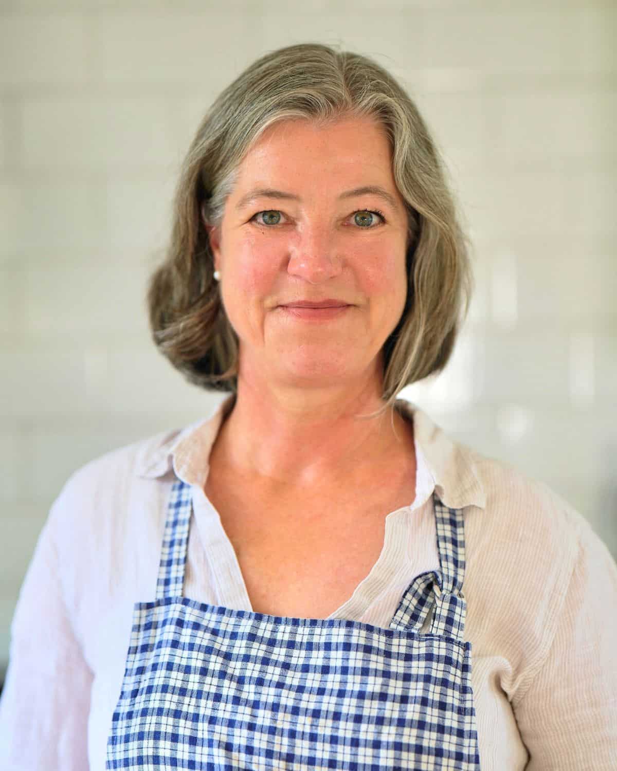 Anja, a middle-aged woman with gray hair wearing a blue and white checkered apron and light-colored shirt, stands in front of a tiled wall, smiling softly—ready to share her simple sourdough recipe.