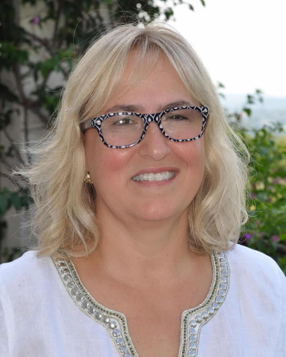 Laura, a woman with shoulder-length blonde hair wearing patterned glasses and a white blouse, smiles outdoors with greenery in the background—radiating warmth reminiscent of Italian hospitality.