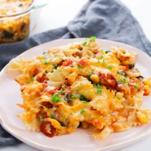 A plate of Enchilada Pasta with melted cheese, vegetables, and herbs, served on a light pink dish with a gray napkin and a glass baking dish in the background.