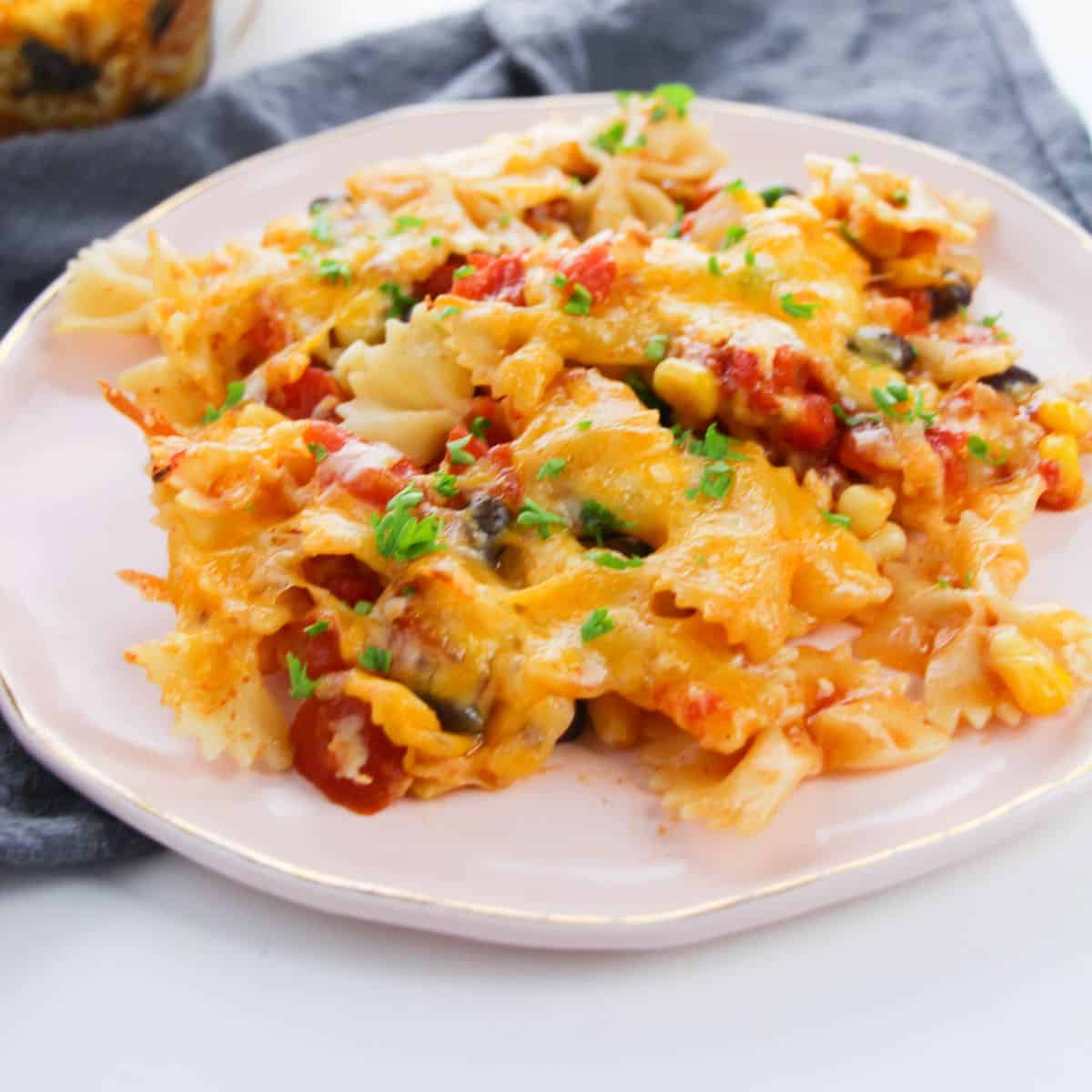 A plate of Enchilada Pasta with bowtie noodles, melted cheese, diced vegetables, and herbs, served on a light pink plate with a gray napkin underneath.