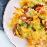 A close-up of cheesy Enchilada Pasta with bow-tie noodles, vegetables, black olives, and zesty tomato sauce, garnished with chopped parsley on a pink plate.