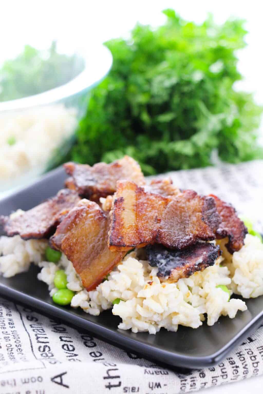 Crispy baked pork belly slices served over rice with green vegetables on a black plate, with fresh parsley in the background.