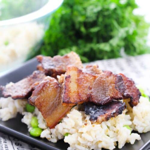 Crispy baked pork belly slices served over rice with green vegetables on a black plate, with fresh parsley in the background.