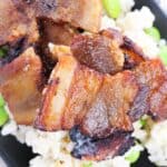 Baked pork belly slices, perfectly crispy, are served on a bed of brown rice and green edamame beans, all elegantly plated on a black dish.