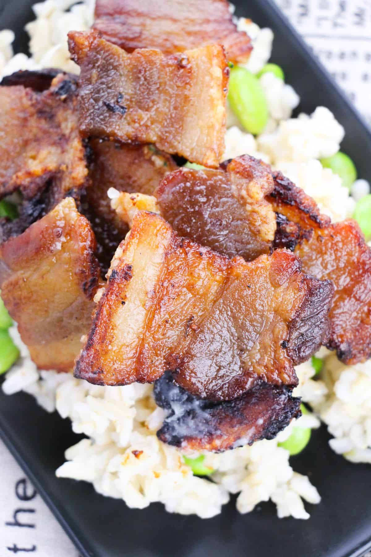 Baked pork belly slices, perfectly crispy, are served on a bed of brown rice and green edamame beans, all elegantly plated on a black dish.