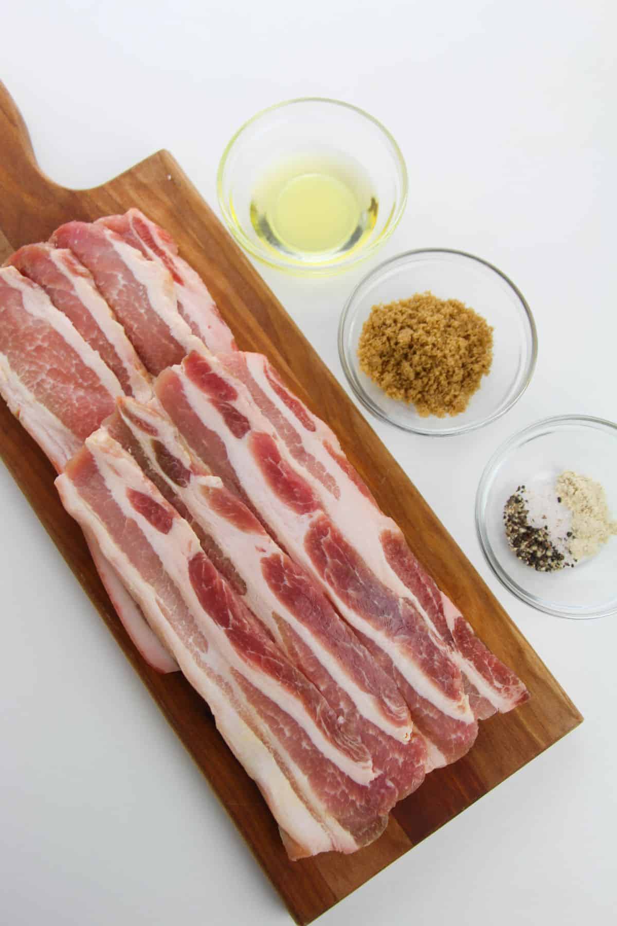 Raw strips on a wooden board, ready to become tender baked pork belly, with bowls of oil, brown sugar, and a salt-pepper mixture arranged on a white surface.