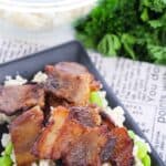 A black plate with slices of baked pork belly on a bed of rice and edamame, topped with parsley, with a bowl of rice in the background.