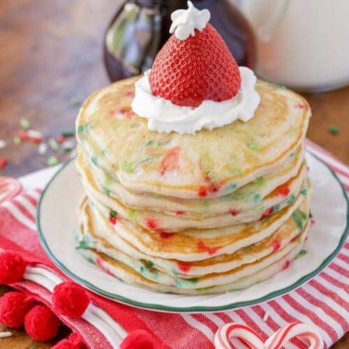 A stack of confetti pancakes topped with whipped cream and a strawberry, served on a plate with a striped napkin, candy canes, and syrup—perfect for Christmas morning breakfast or adding to your collection of cozy breakfast recipes.