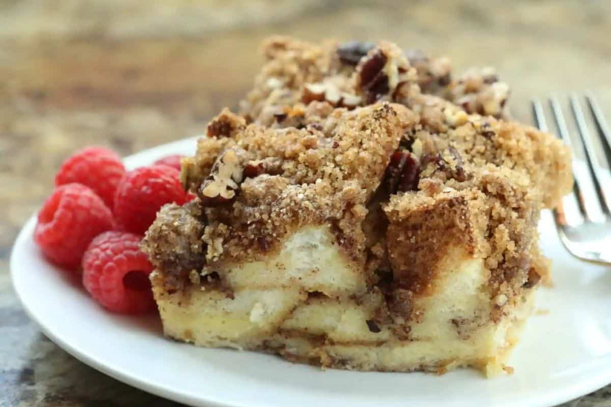 A slice of French toast casserole with crumb topping and pecans is served on a white plate with fresh raspberries and a fork—perfect for cozy breakfast recipes or as a delicious Christmas morning breakfast.
