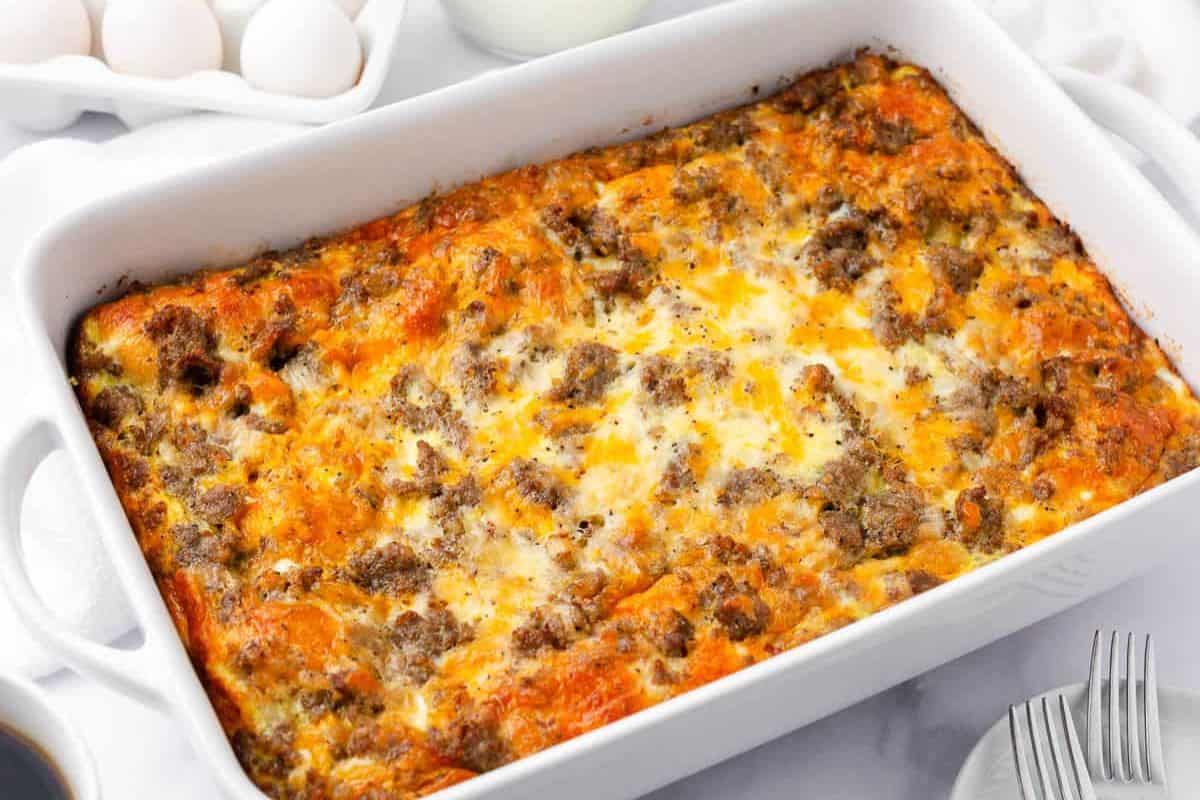 A baked breakfast casserole with browned sausage, melted cheese, and eggs in a white rectangular dish—perfect for cozy Christmas breakfast gatherings, surrounded by fresh eggs and serving utensils.